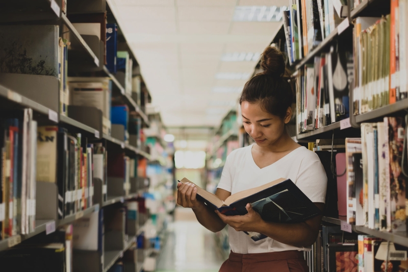 Female college student reading book in the library aisle